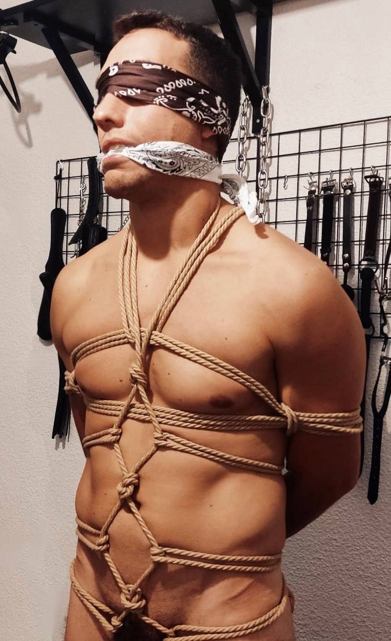 A muscular man is restrained in a complex rope harness, blindfolded, and gagged in a dungeon setting.