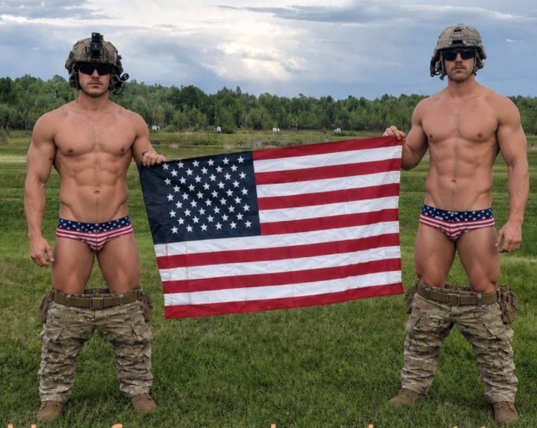 Two muscular men in tactical helmets and American flag briefs hold a flag in an outdoor field with their camo pants dropped around their ankles.