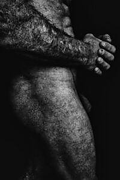 A black and white profile photograph of an extremely hairy, muscular man standing against a dark background.
