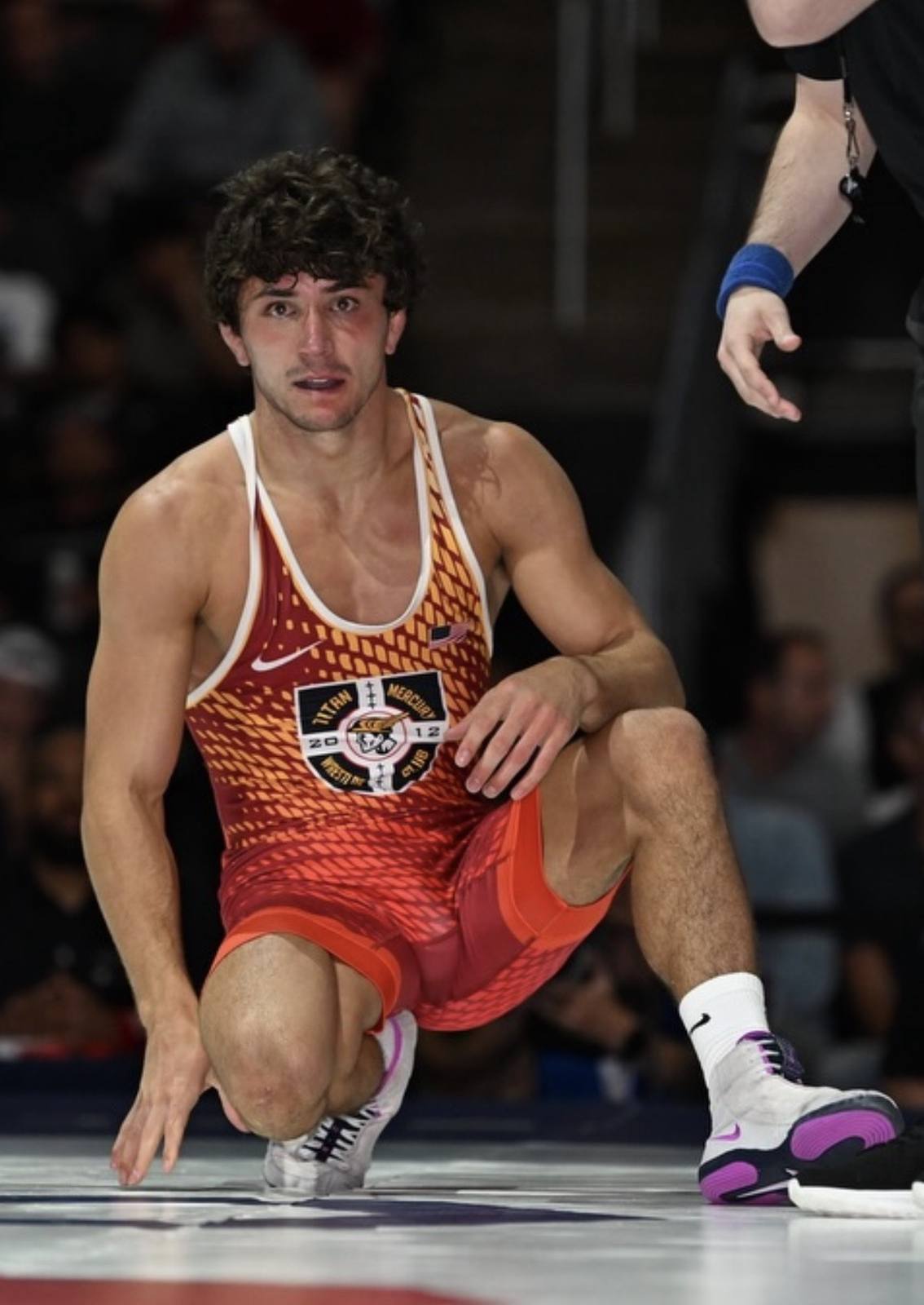 A young athletic man in a patterned red wrestling singlet kneels on a competition mat.
