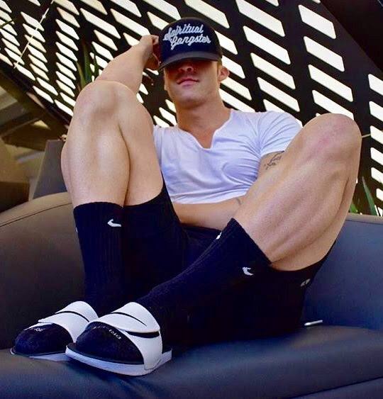 A muscular jock sits back on a sofa wearing a white t-shirt, gym shorts, black crew socks, and slides.