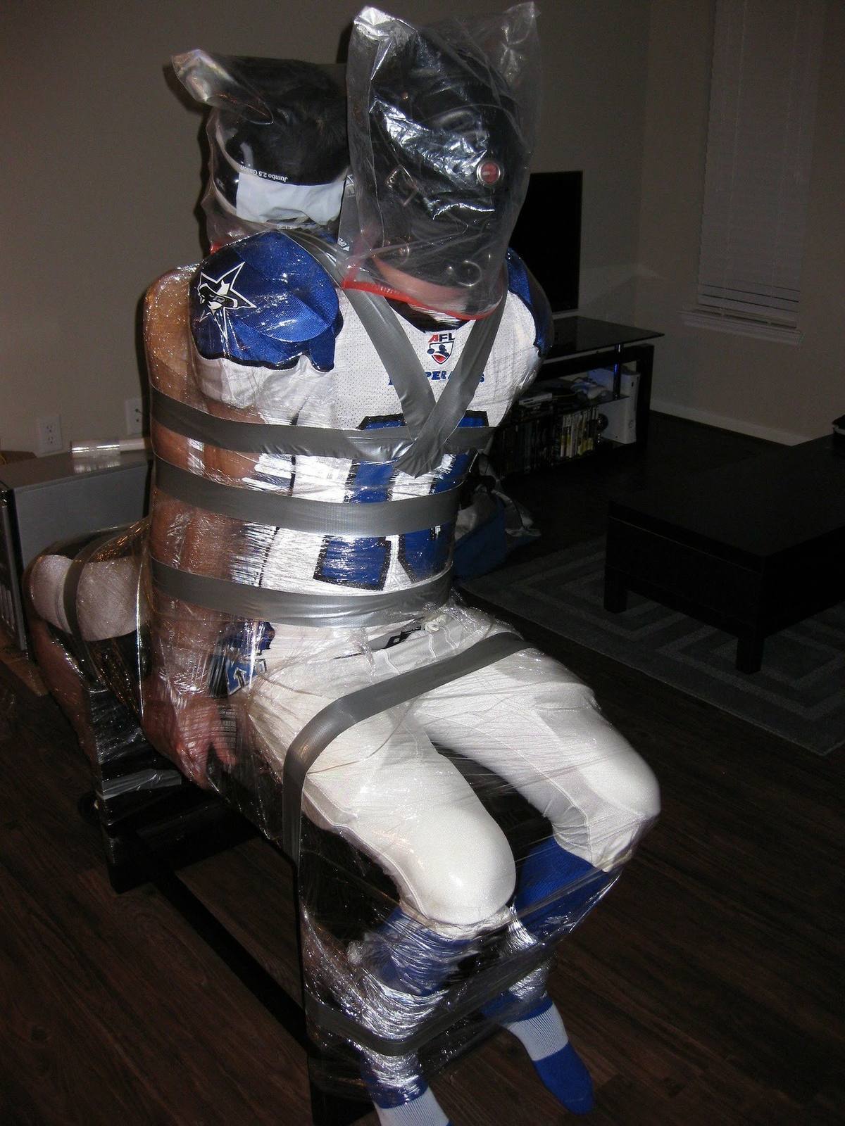 A person in a full American football kit and leather hood is heavily bound with plastic wrap and duct tape, including a plastic bag over their head.