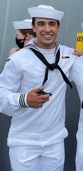 A fit young man smiles while wearing a traditional white US Navy sailor uniform and hat.