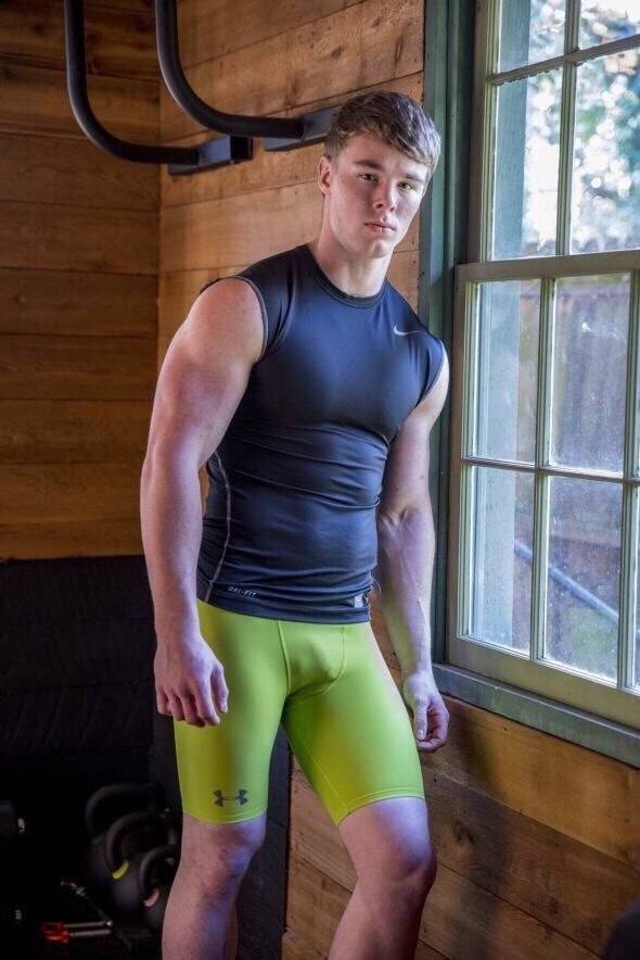 A muscular young man in a black compression top and tight lime green spandex shorts poses in a rustic home gym near a window.