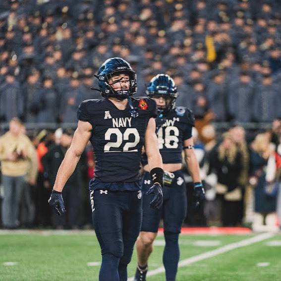 Two muscular athletes in full Navy football kits walk on the grass field in front of a stadium crowd.