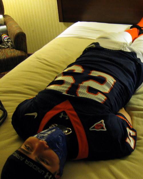 A man wearing a full football uniform is restrained on a bed with a tape gag and collar.