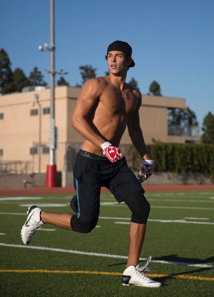 A shirtless twunk in gym shorts and football gloves runs across an outdoor turf field.