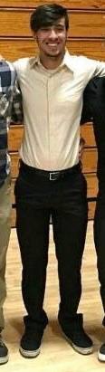 A smiling twink wearing a tan button-up shirt and black trousers stands in a gym.