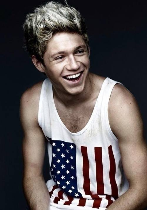 A young man with blonde highlights and a hairy chest smiles while wearing a white tank top featuring an American flag print.