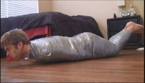 A man is completely cocooned in silver duct tape from neck to feet with a red ball gag in his mouth, lying prone on a bedroom floor.