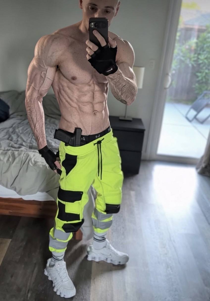 A shirtless, shredded muscle stud takes a mirror selfie in a bedroom wearing high-visibility cargo pants and fingerless gloves.