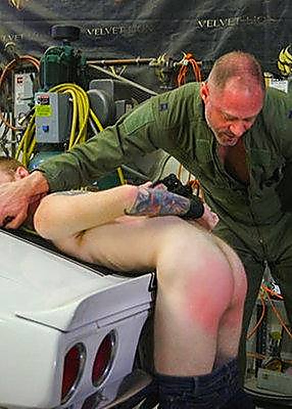 A mature man in a military flight suit spanks a bound man bent over the back of a car in a garage.