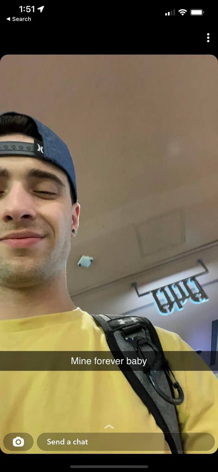A young, smiling man with stubble wears a yellow t-shirt and a backwards blue baseball cap while carrying a backpack in a public indoor setting.