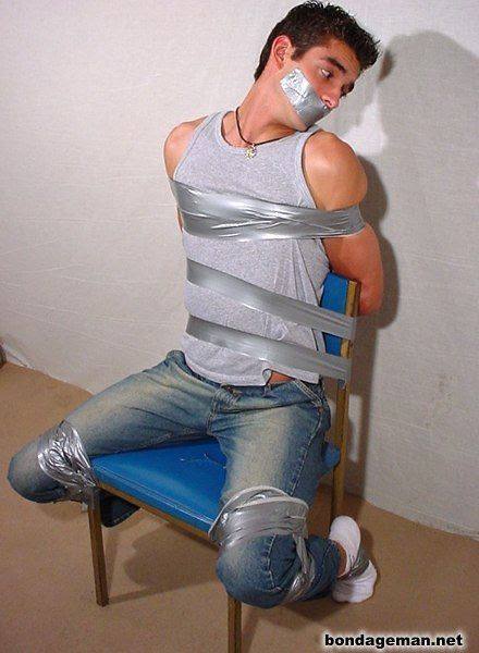 A young athletic man in a tank top and jeans is gagged and bound to a chair with silver duct tape.