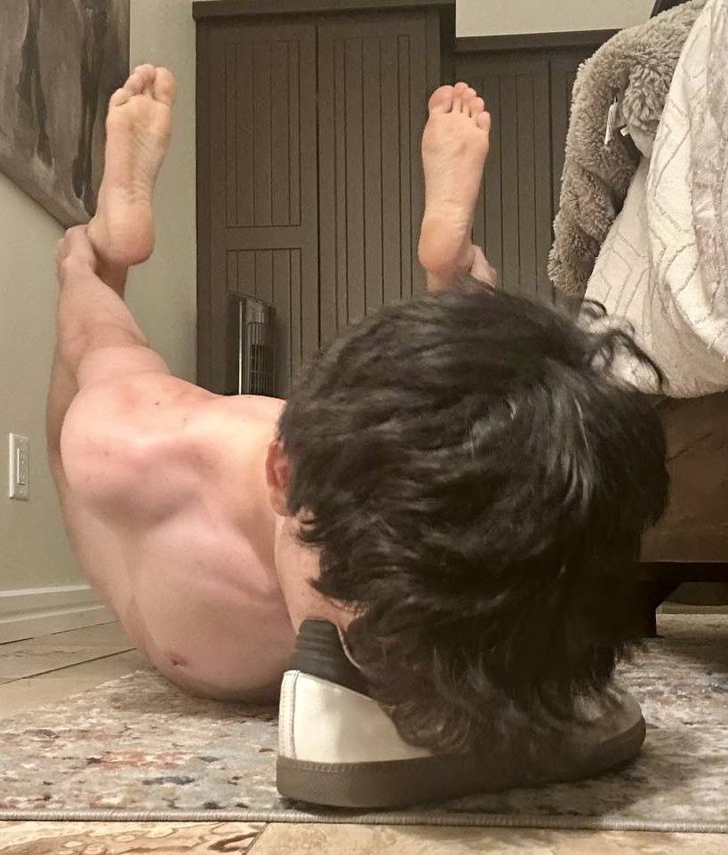 A shirtless young man lies prone on a bedroom rug, resting his head on a white sneaker while holding his bare feet in the air.