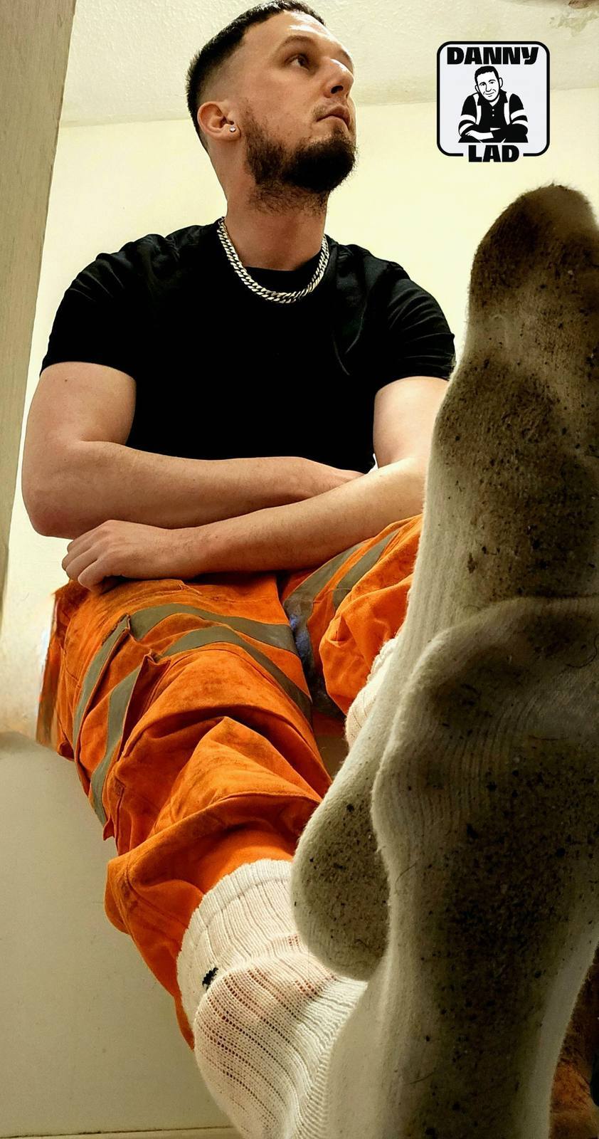 A bearded man in orange high-visibility construction pants sits with his dirty white socks prominently displayed in a POV perspective.