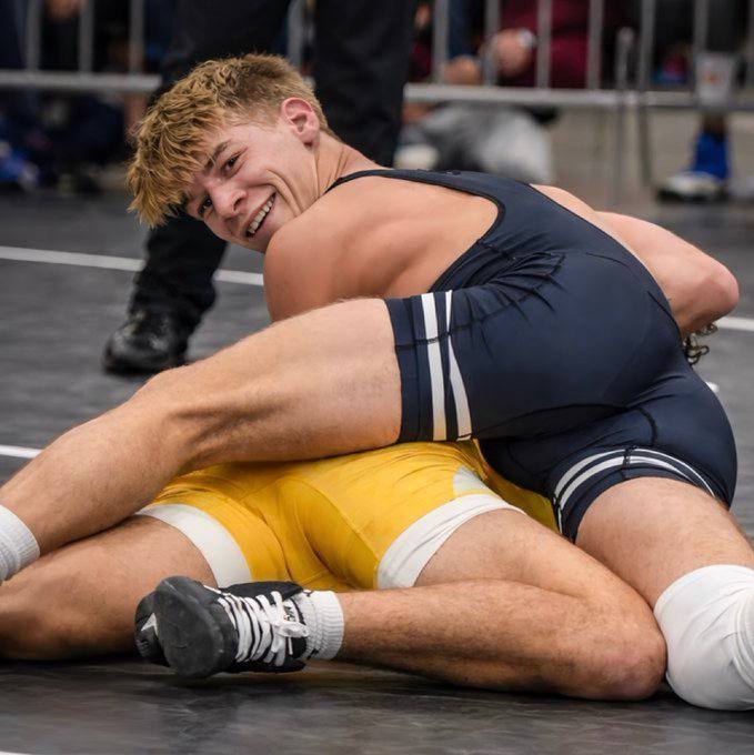 Two athletic young men in lycra wrestling singlets engage in a grappling match on a gym mat.