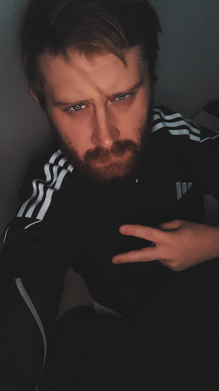 A bearded man in a black Adidas tracksuit top poses for a selfie.