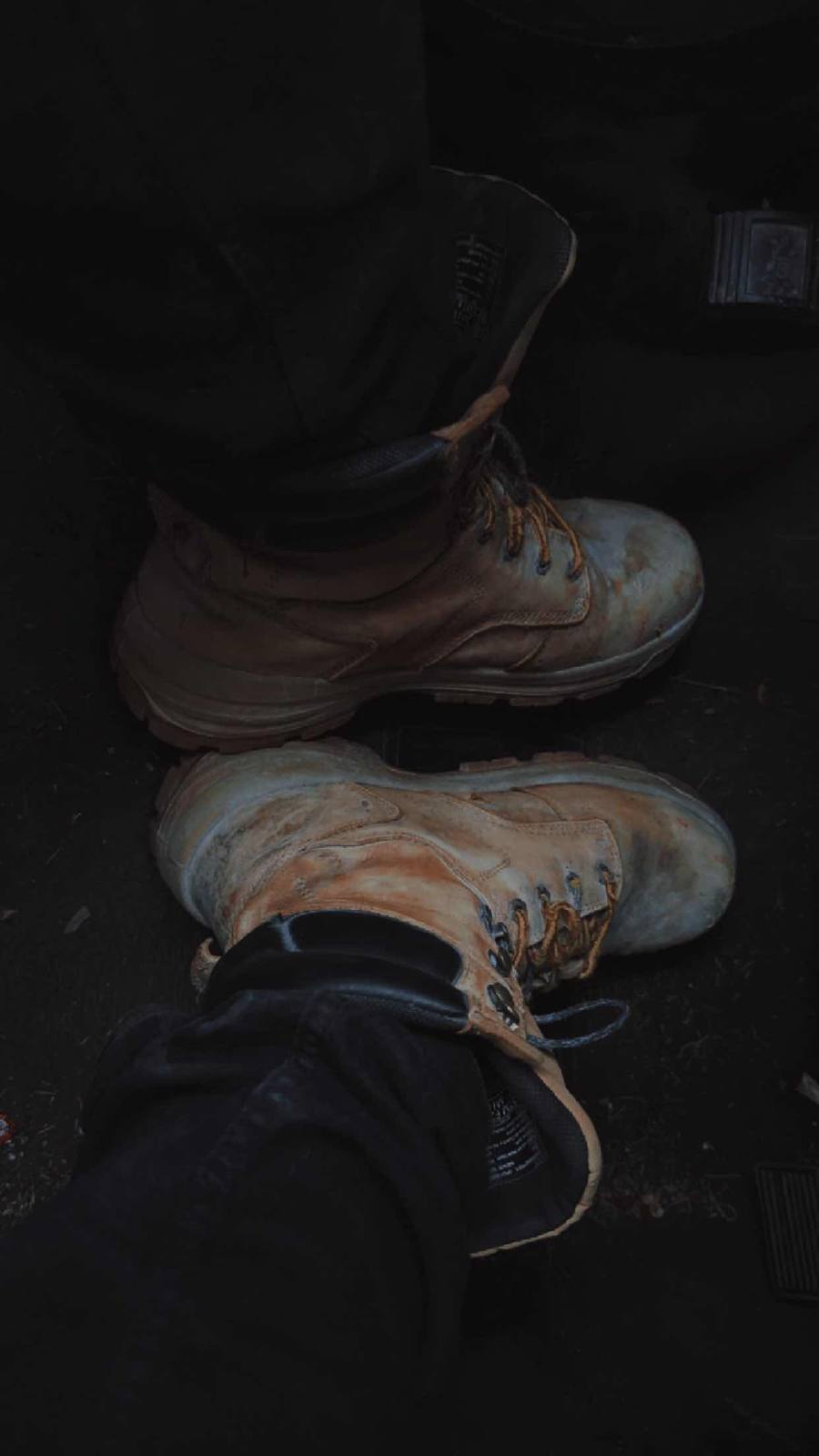 A POV shot looks down at a pair of worn, dirty tan leather work boots resting on a dark vehicle floor.