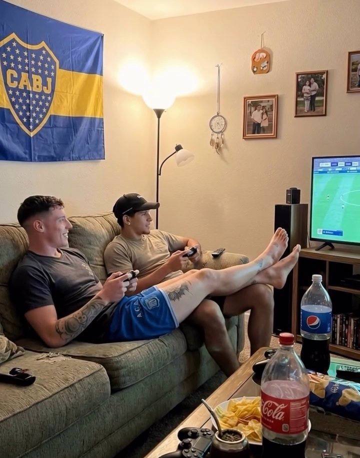 Two men in athletic t-shirts and shorts relax on a couch playing video games with their bare feet on display.