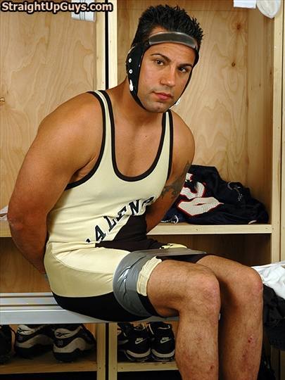 A muscular man in a wrestling singlet and headgear sits in a locker room with his arms behind his back and his thighs bound with duct tape.
