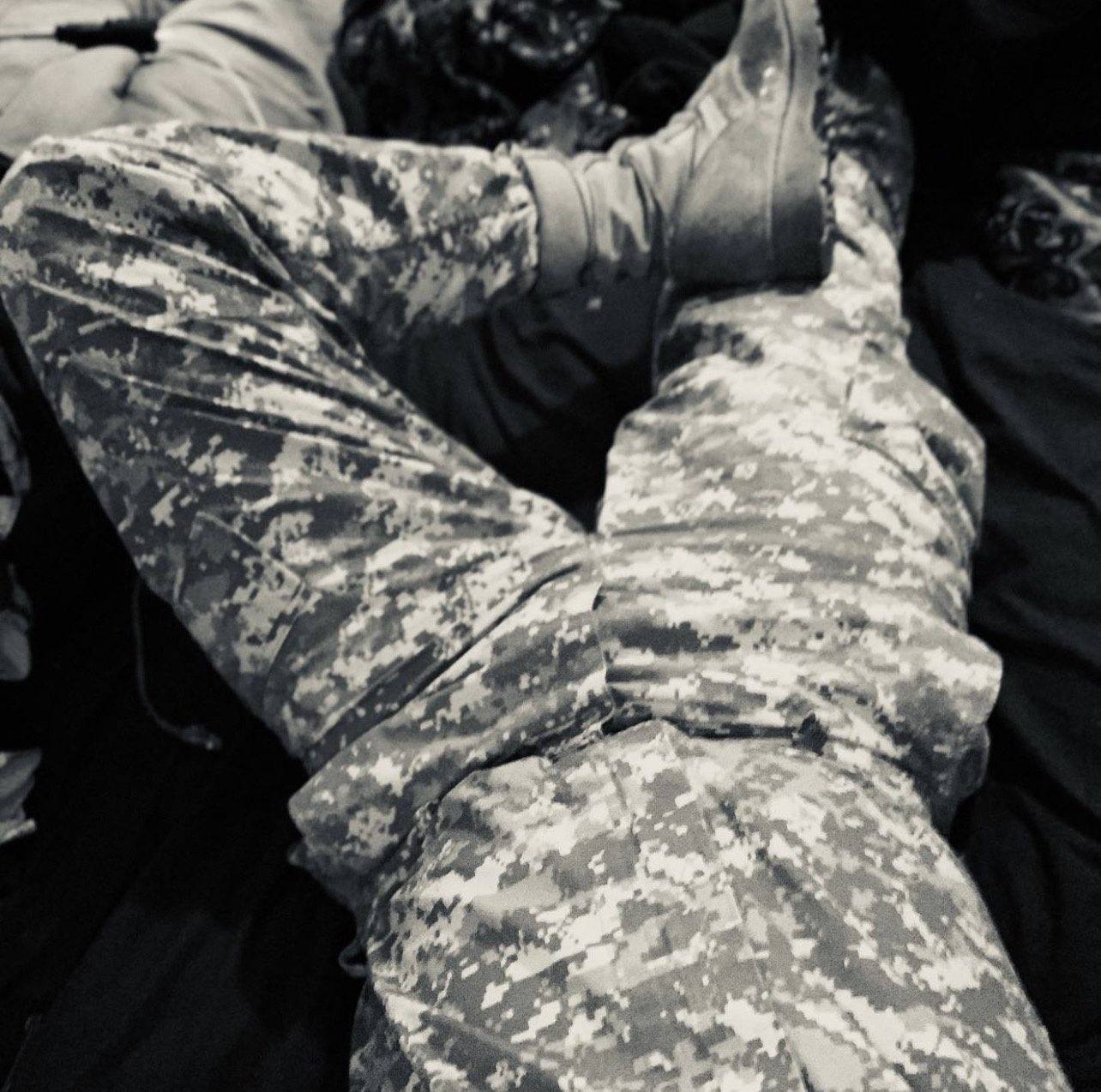 A man lies on a bed wearing digital camouflage trousers and combat boots.