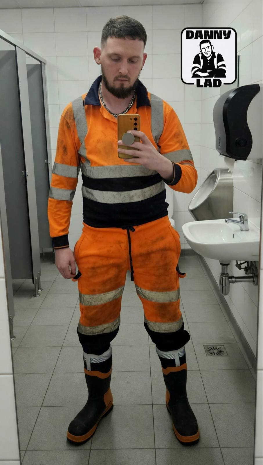 A bearded man in dirty high-visibility construction workwear and rubber boots takes a mirror selfie in a public restroom.