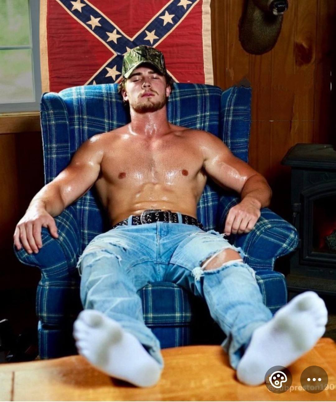 A sweaty, muscular man in a camo cap and ripped jeans relaxes in an armchair with his white-socked feet propped up.