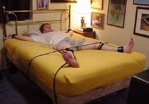 A man is restrained in a spread-eagle position on a bed using leather cuffs and rope attached to the bed frame.
