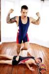 A muscular tattooed man in a blue and red wrestling singlet flexes triumphantly while standing with one foot on his defeated opponent.