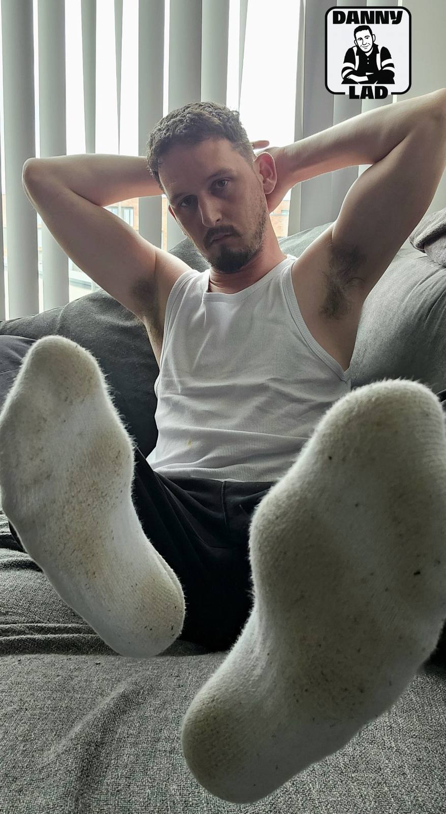 A bearded man in a white tank top sits on a couch with his hands behind his head, prominently displaying the soles of his dirty white socks to the camera.
