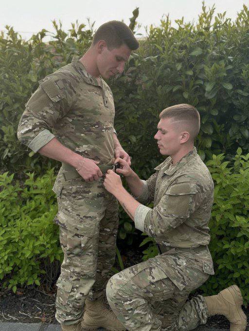 A man in a camouflage military uniform kneels to adjust the belt of another uniformed man standing before him in an outdoor setting.