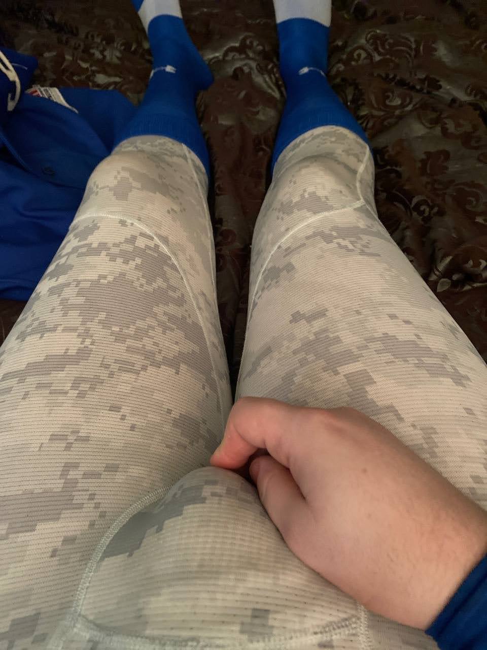 A POV shot looking down at a man wearing digital camo compression leggings and blue athletic socks, with his hand resting on his crotch.