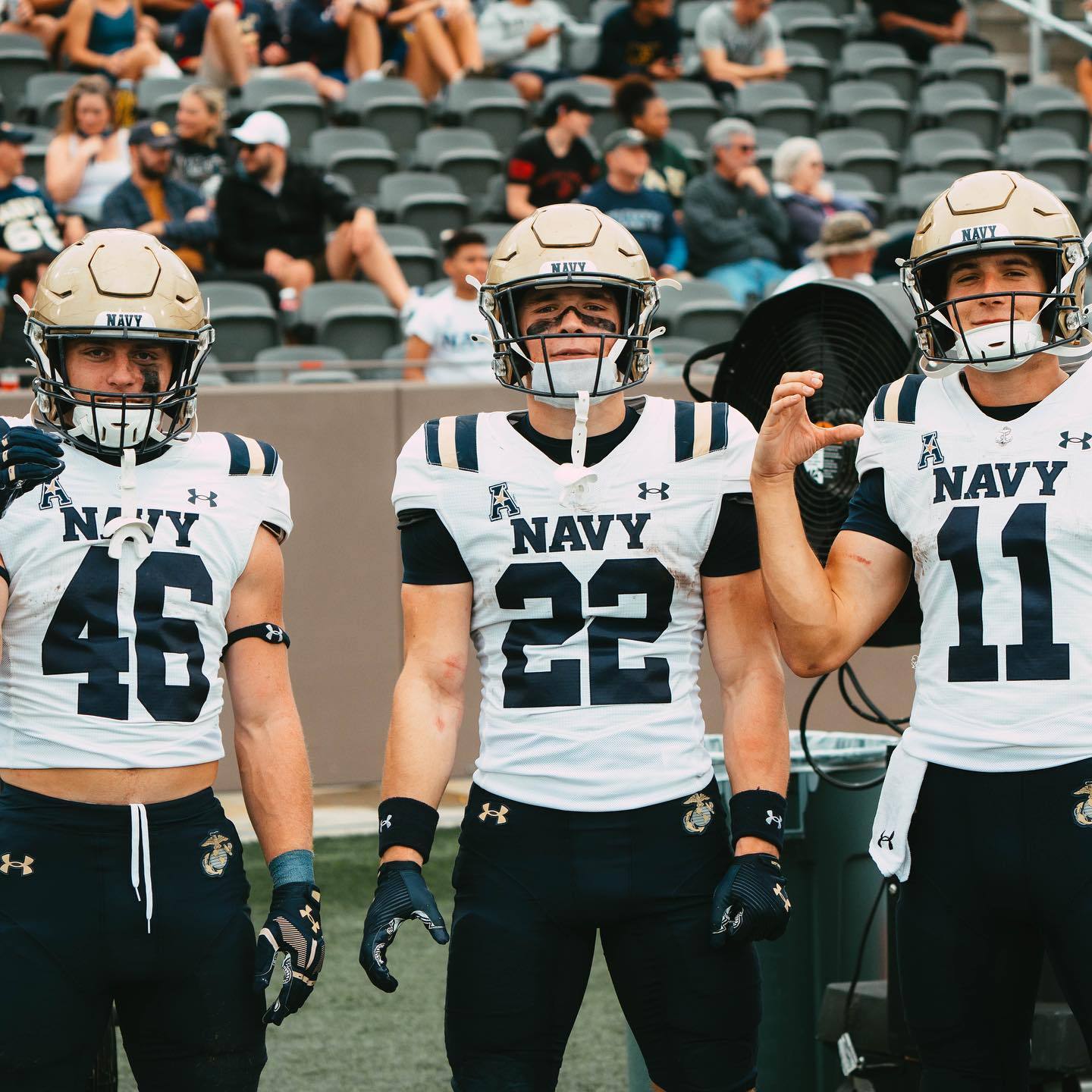 Three muscular football players in Navy uniforms and helmets stand on a stadium sideline.