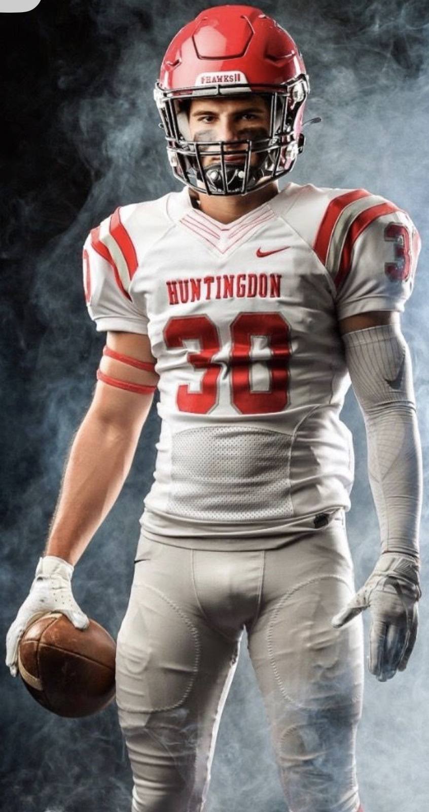 A muscular jock poses in a full Huntingdon football uniform, including a red helmet and tight white pants that highlight his build.