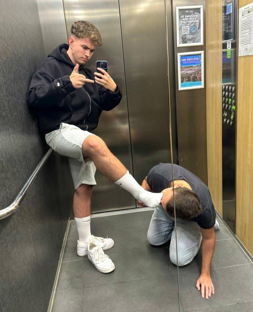 A young man takes a mirror selfie in an elevator while holding his socked foot up for another kneeling man to worship.