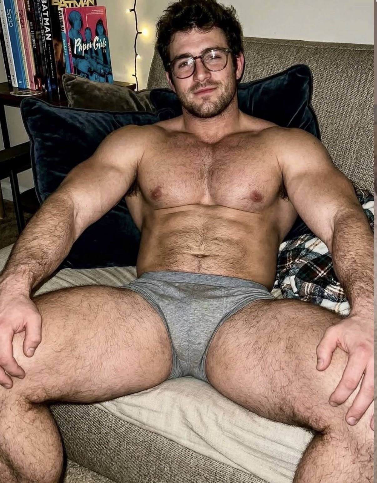 A muscular, bearded man with glasses and a hairy chest poses sitting on a couch wearing grey cotton briefs.