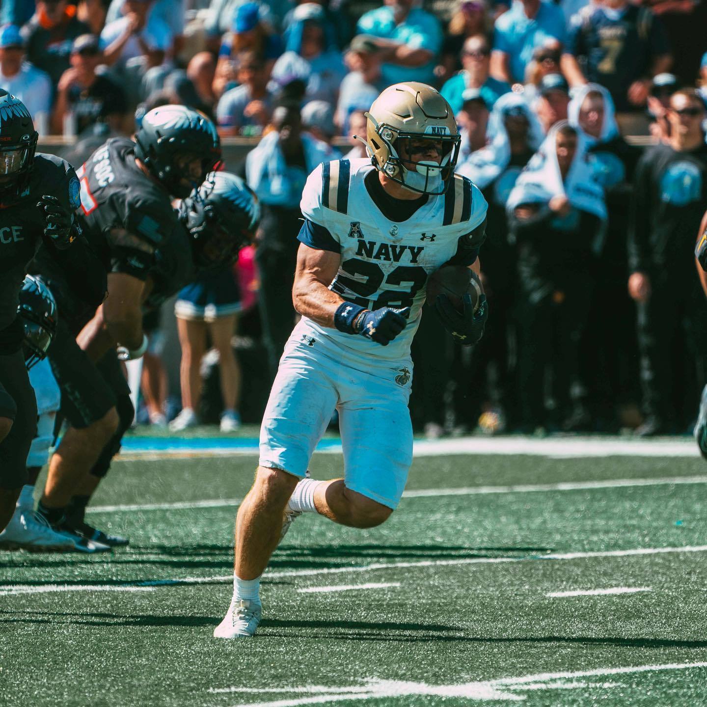 A muscular football player in a white and gold Navy uniform runs across the field during a game.