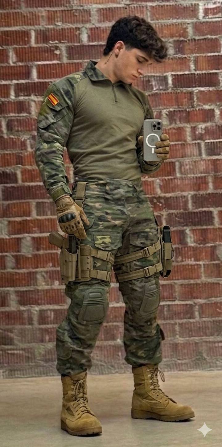 A muscular young man in full military camo and tactical gear takes a selfie against a brick wall.