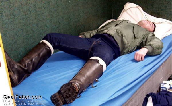 A man in a military flight jacket and denim jeans lies on a bed wearing tall leather boots.