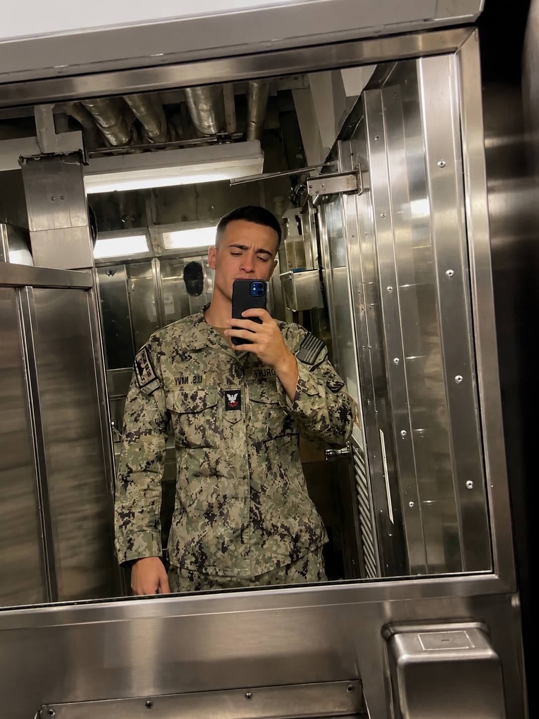 A young man with a buzzcut wears a US Navy camouflage uniform for a mirror selfie in an industrial locker room.