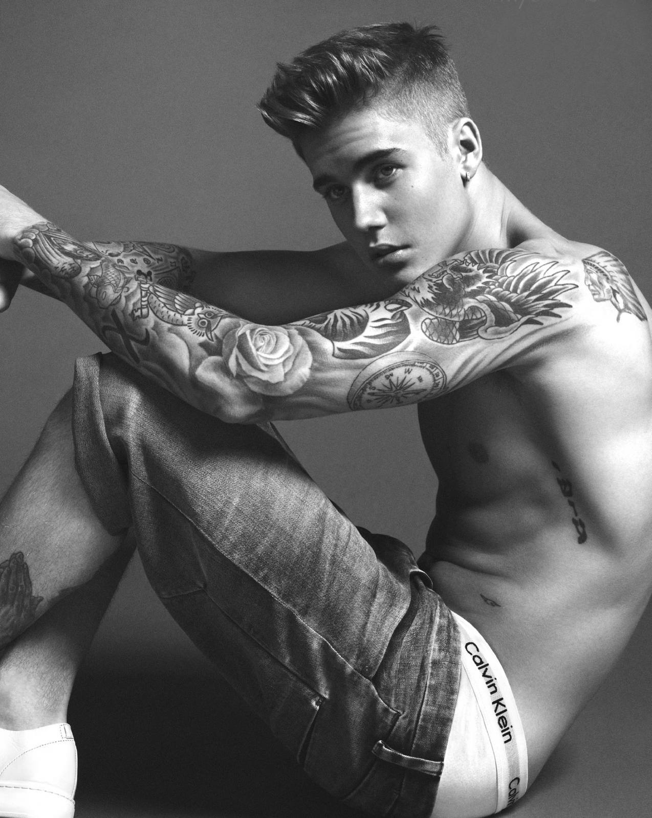 A shirtless twunk with extensive tattoos on his arms and torso poses in cuffed denim shorts and white underwear.