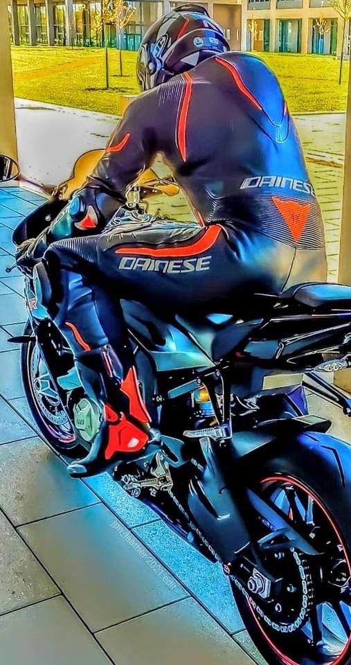 A person clad in a black and red Dainese leather racing suit sits atop a motorcycle outdoors.
