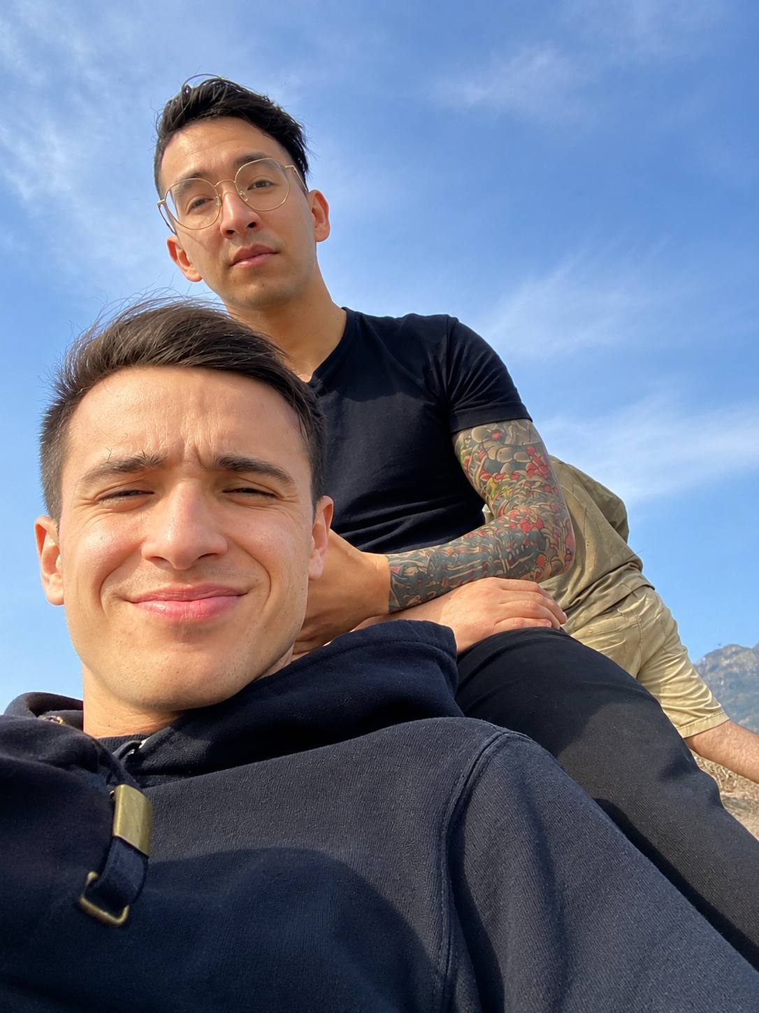 Two clean-shaven men, one with a full sleeve tattoo, pose for a selfie while outdoors in a mountainous area.