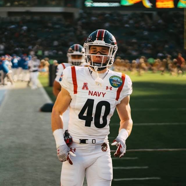 A muscular young football player in a full Navy uniform stands on the field wearing his helmet and gloves.