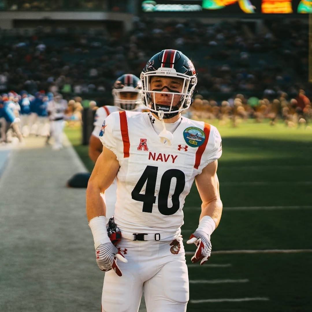 A muscular young man in a full Navy football uniform stands on a stadium field.
