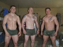 Three shirtless, muscular men in short olive green military-style gym shorts pose for a photo in a garage.