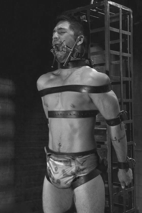 A muscular submissive man is restrained with leather straps, a collar, and a mouth spreader while standing in a dungeon setting.
