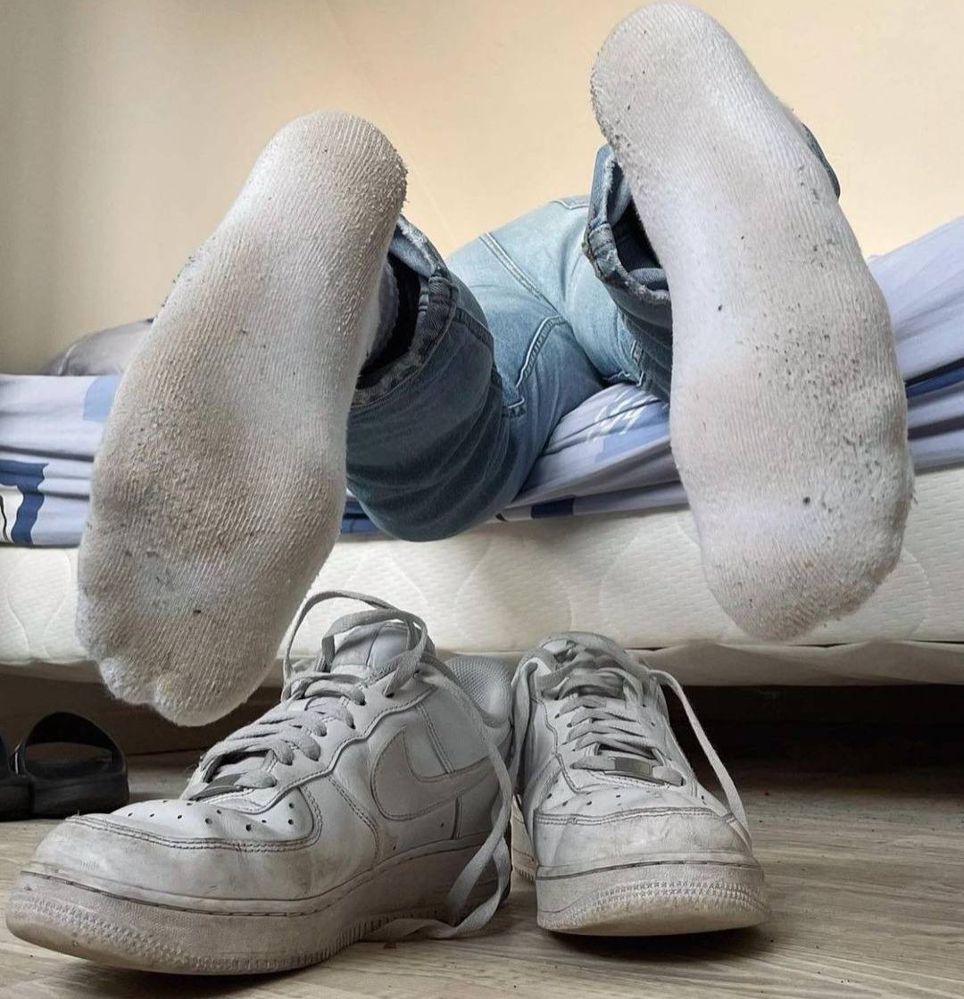 A person wearing jeans and dirty white socks rests with their feet up, next to their dirty white sneakers on the floor.