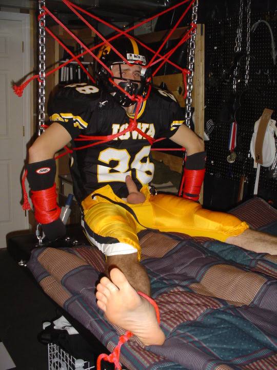 A man wearing a full football uniform is restrained by intricate red rope bondage in a dungeon setting with his genitals exposed.